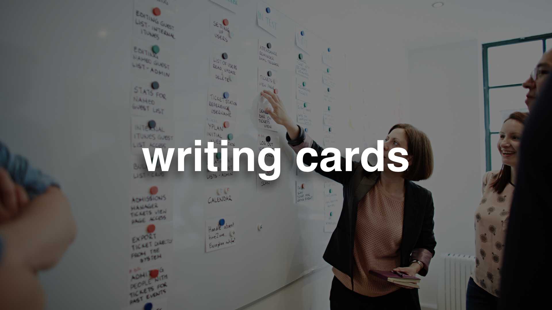 A photo of some people standing in front of an agile wall with index cards on it - text: writing cards